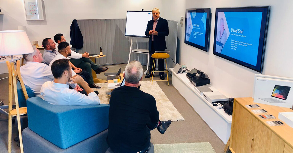 news - a Office 365 consultant standing by a Microsoft Surface Hub, presenting to a group of people sitting down